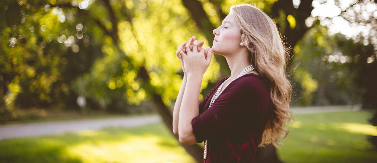 A blonde woman praying with spring trees in the background