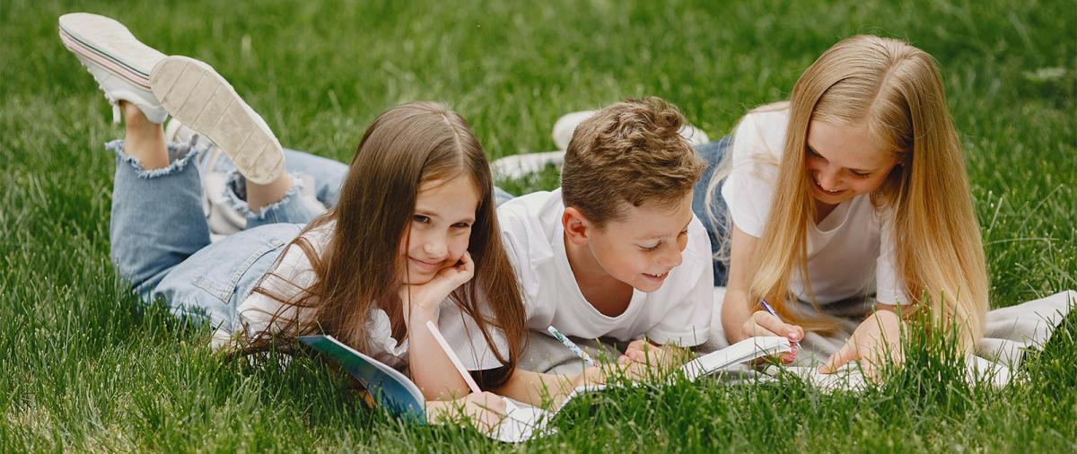 Three young children laying in a grassy area reading.